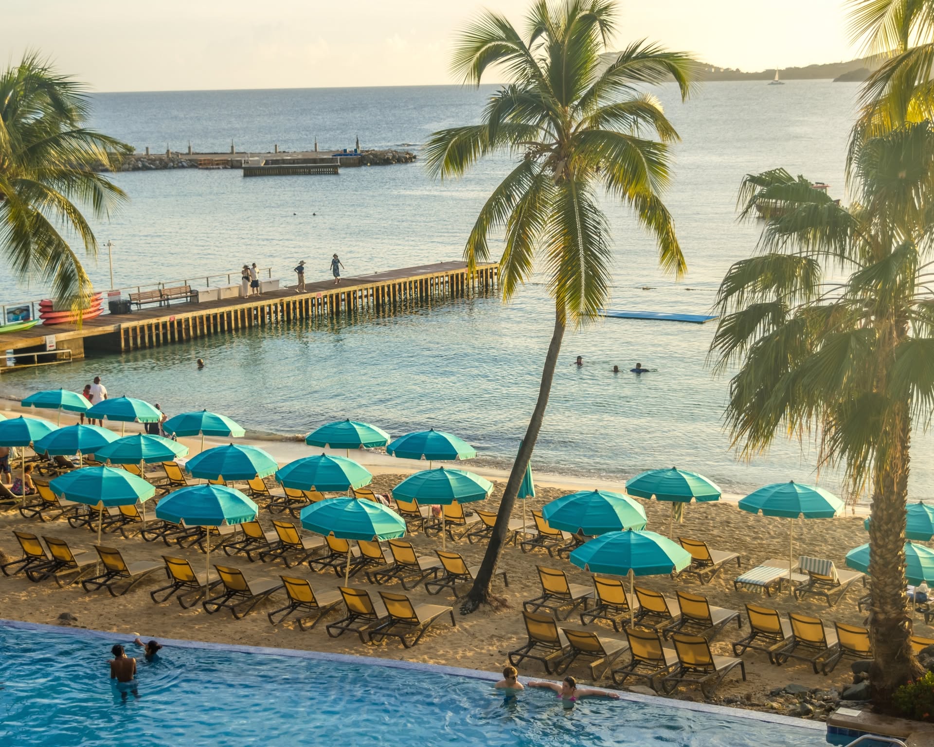 Beach with palm trees, turquoise umbrellas, lounge chairs, and a pier extending into the sea.
