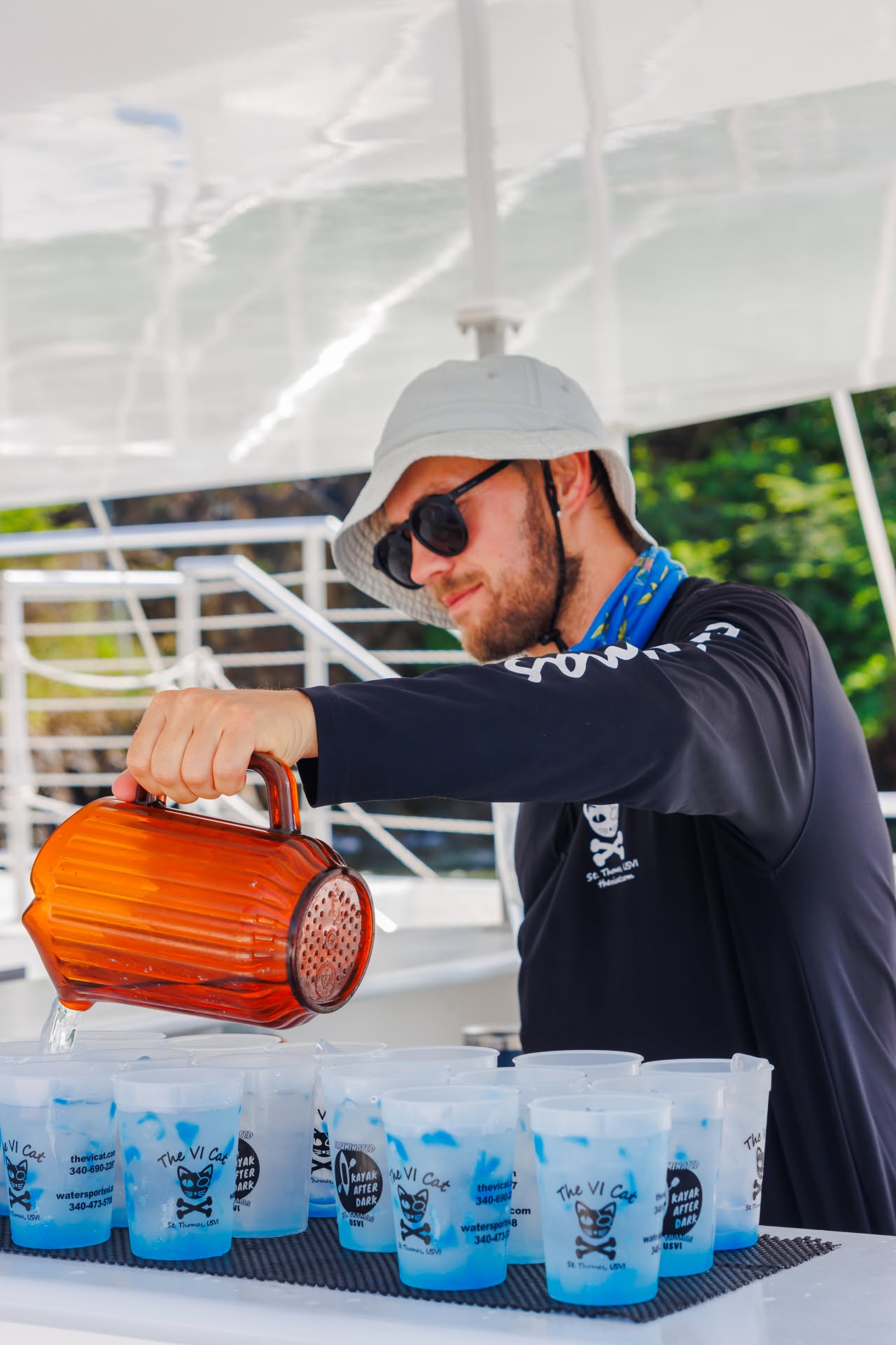 Man in hat pouring drinks from a brown jug into plastic cups on a boat.