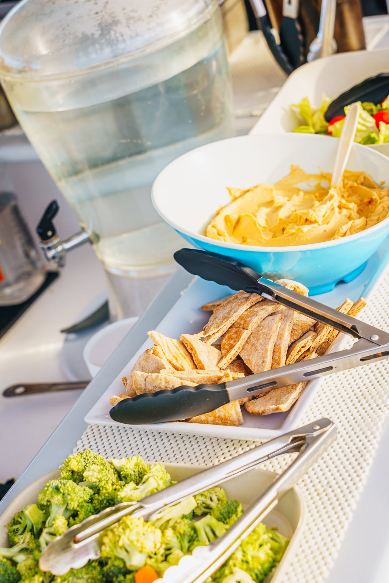 Buffet with chips, pita, broccoli, and salad beside a water dispenser.