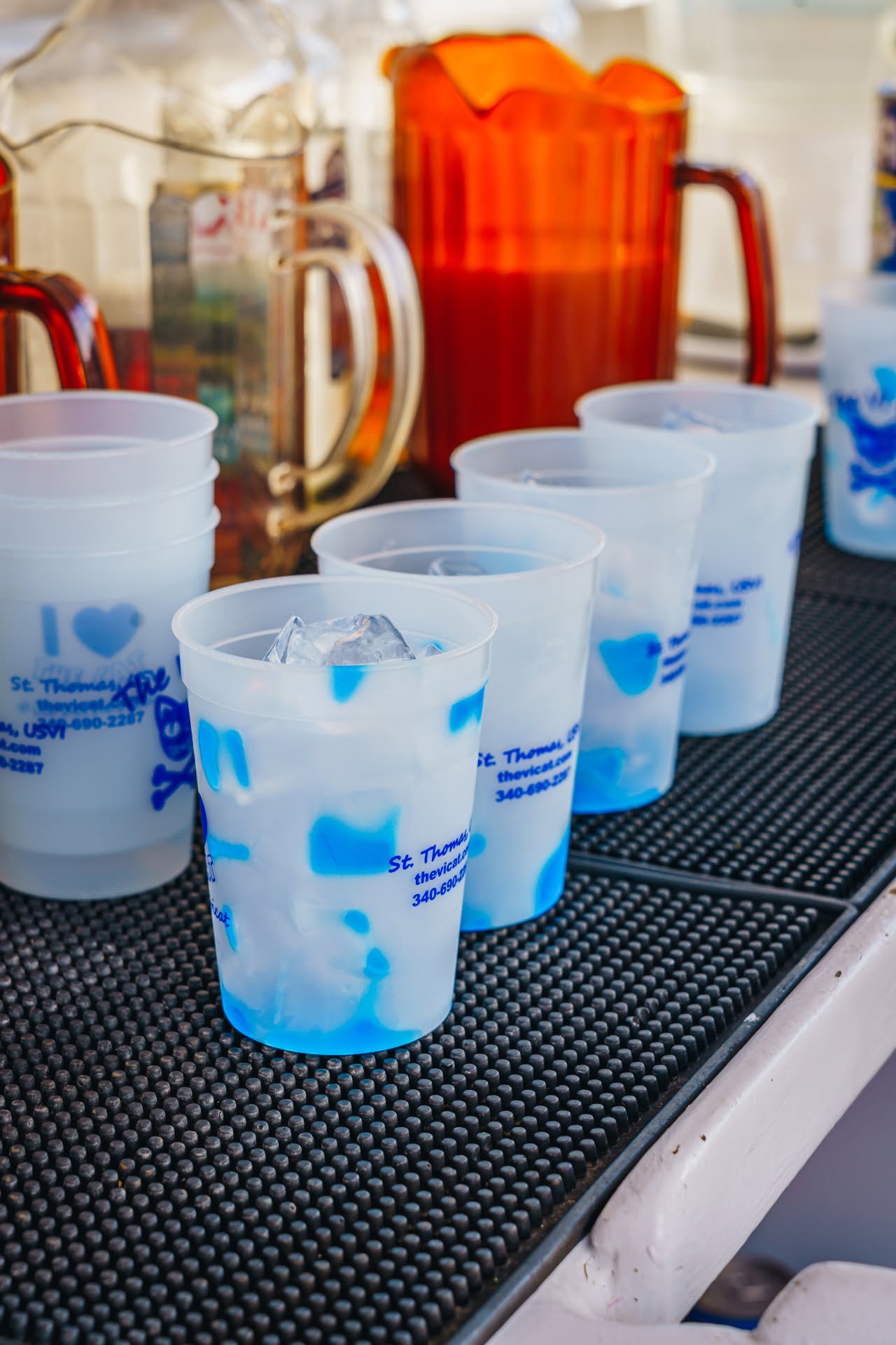 Plastic cups with ice lined up on a bar mat, a jug of orange liquid behind.