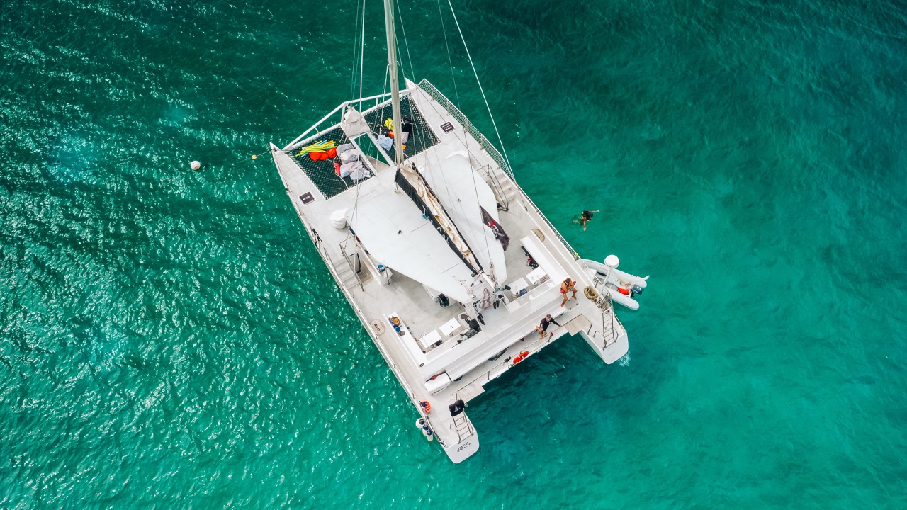 Aerial view of a catamaran with white sails on clear turquoise water.