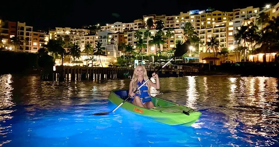 Person kayaking at night on glowing blue water near illuminated buildings.