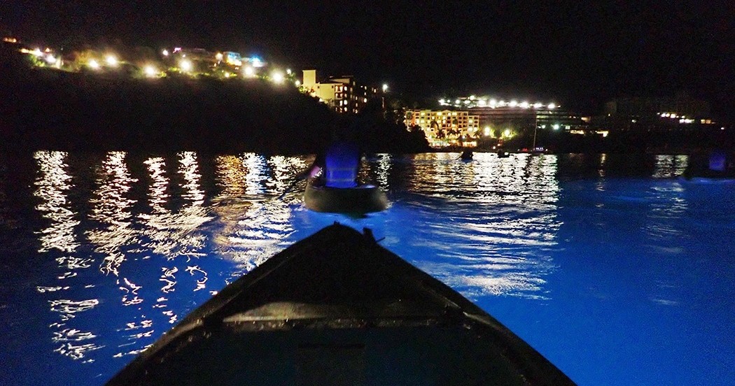Boat on glowing blue water at night with city lights in background.