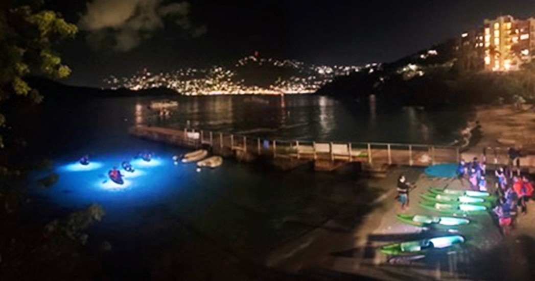 Nighttime scene with kayaks on beach, glowing water, and lit hills.