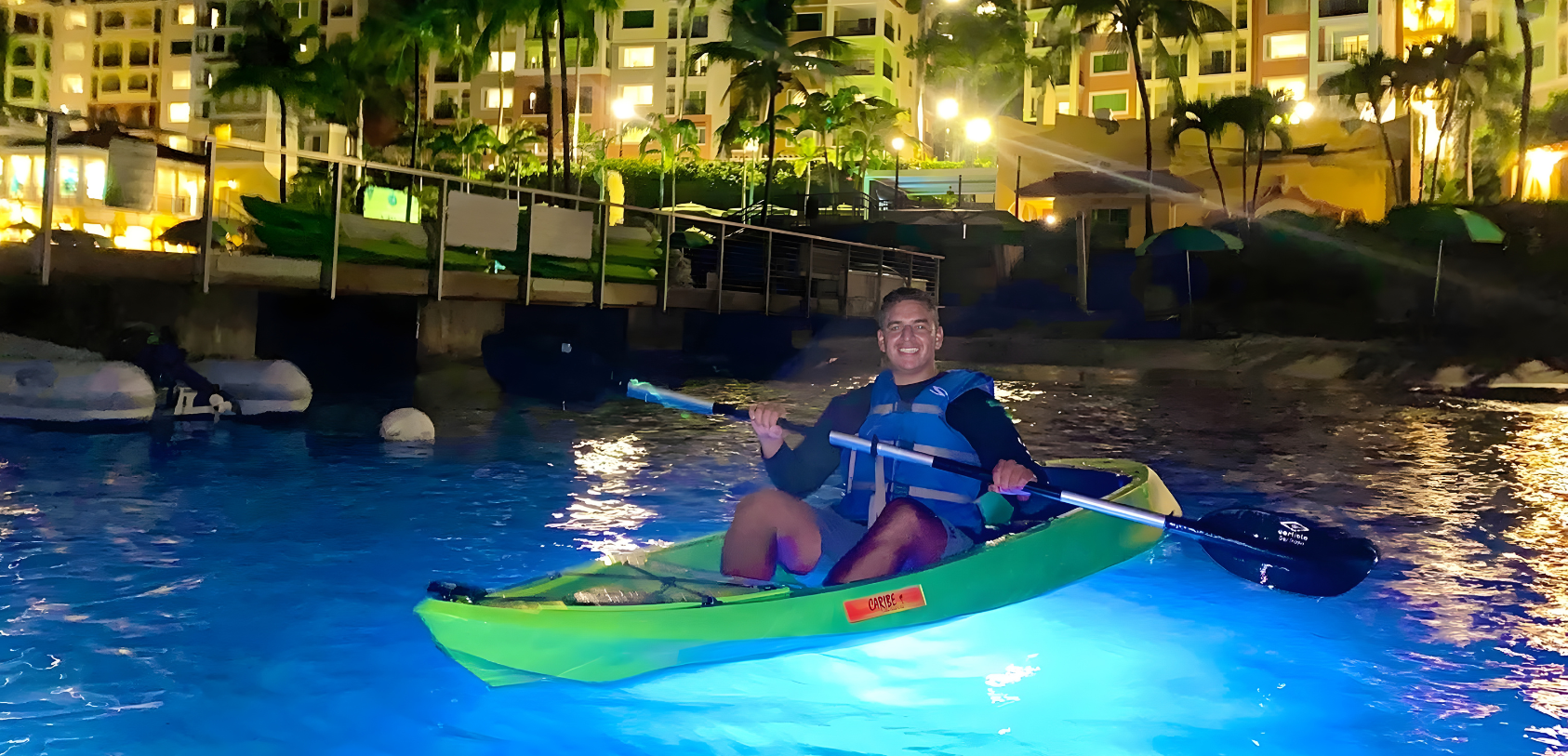 Person kayaking on illuminated water at night near brightly lit buildings.