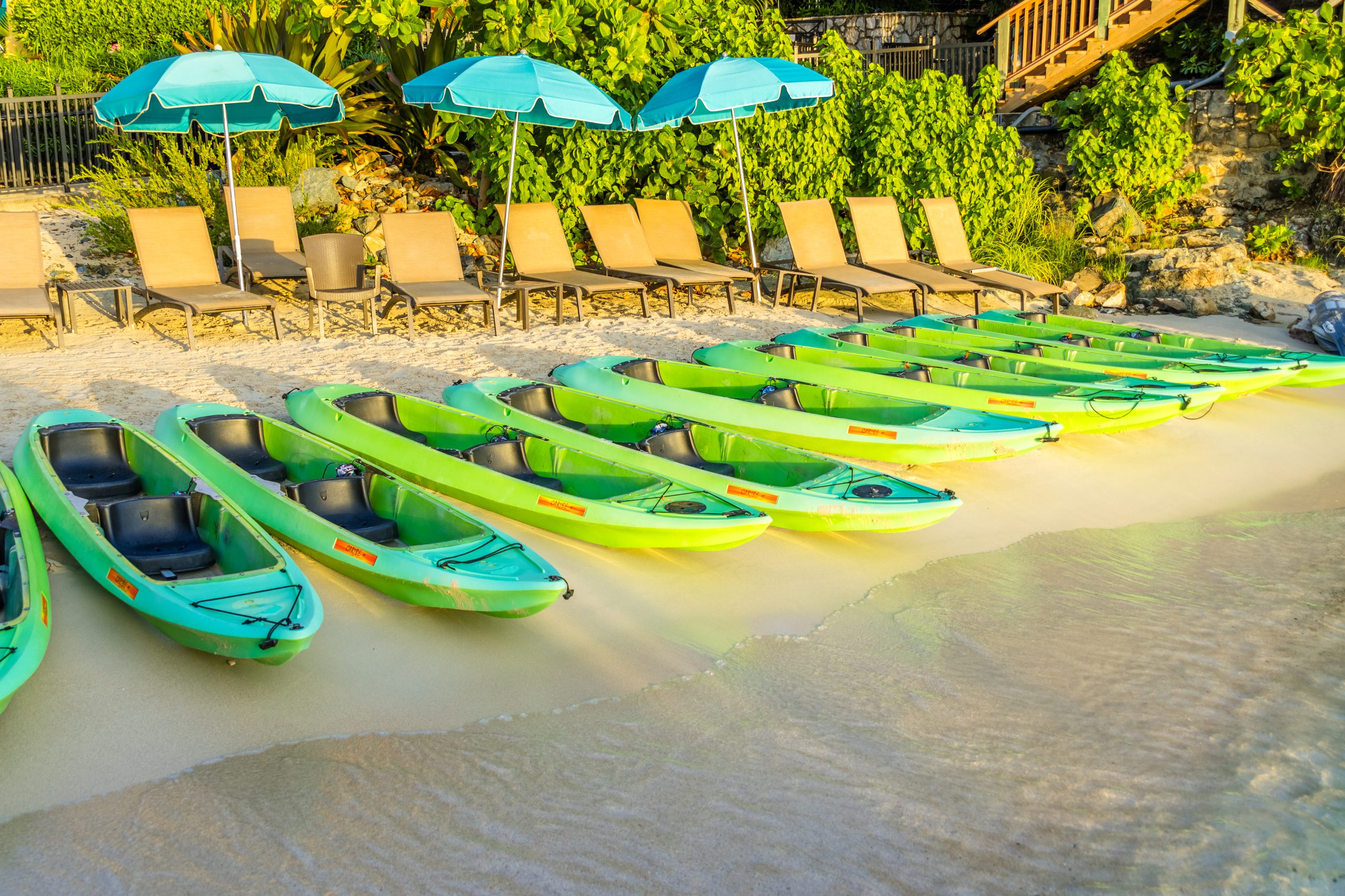 Line of green kayaks on sandy beach near sun loungers and blue umbrellas.