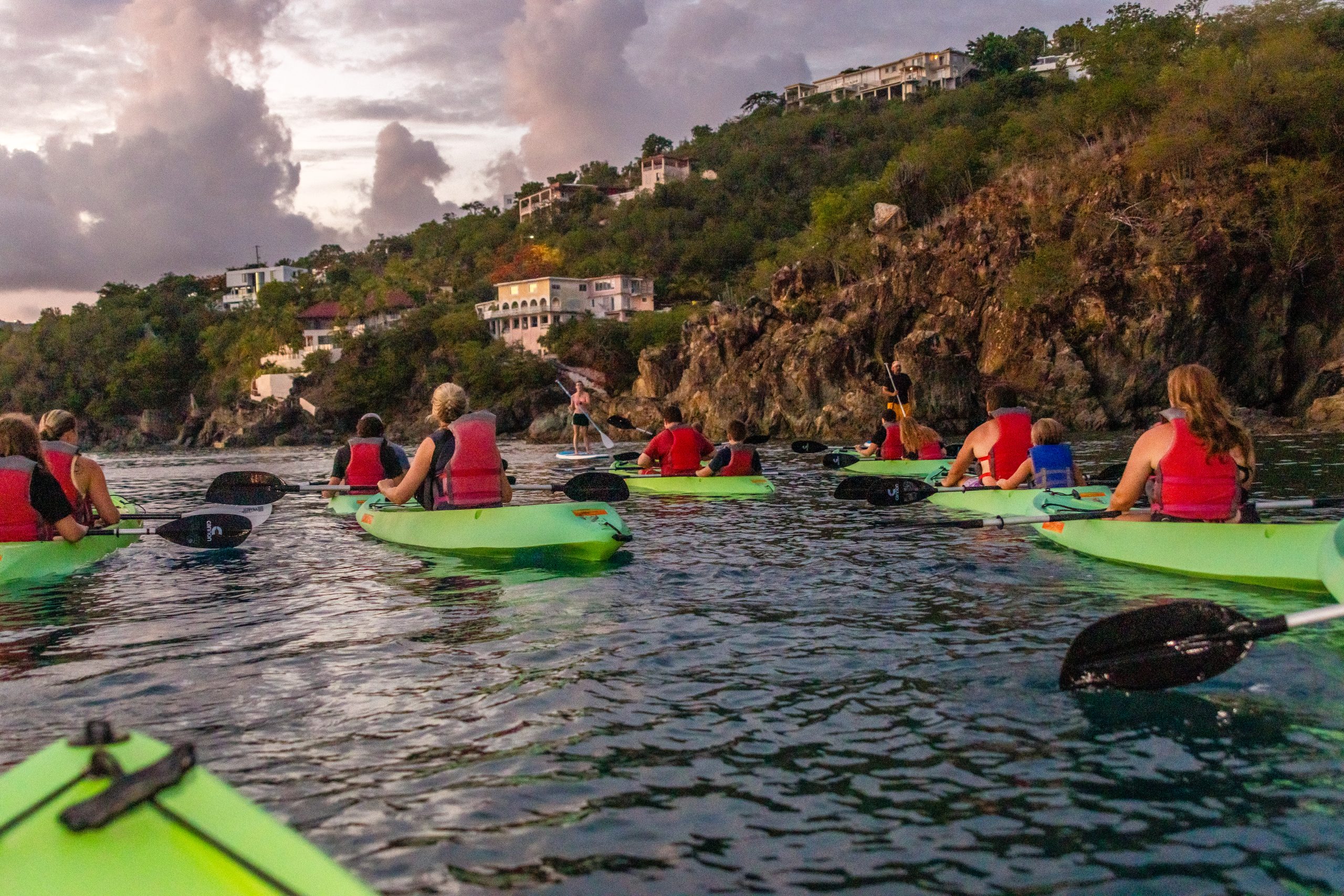 Group kayaking near rocky shore with houses on hillside at sunset.