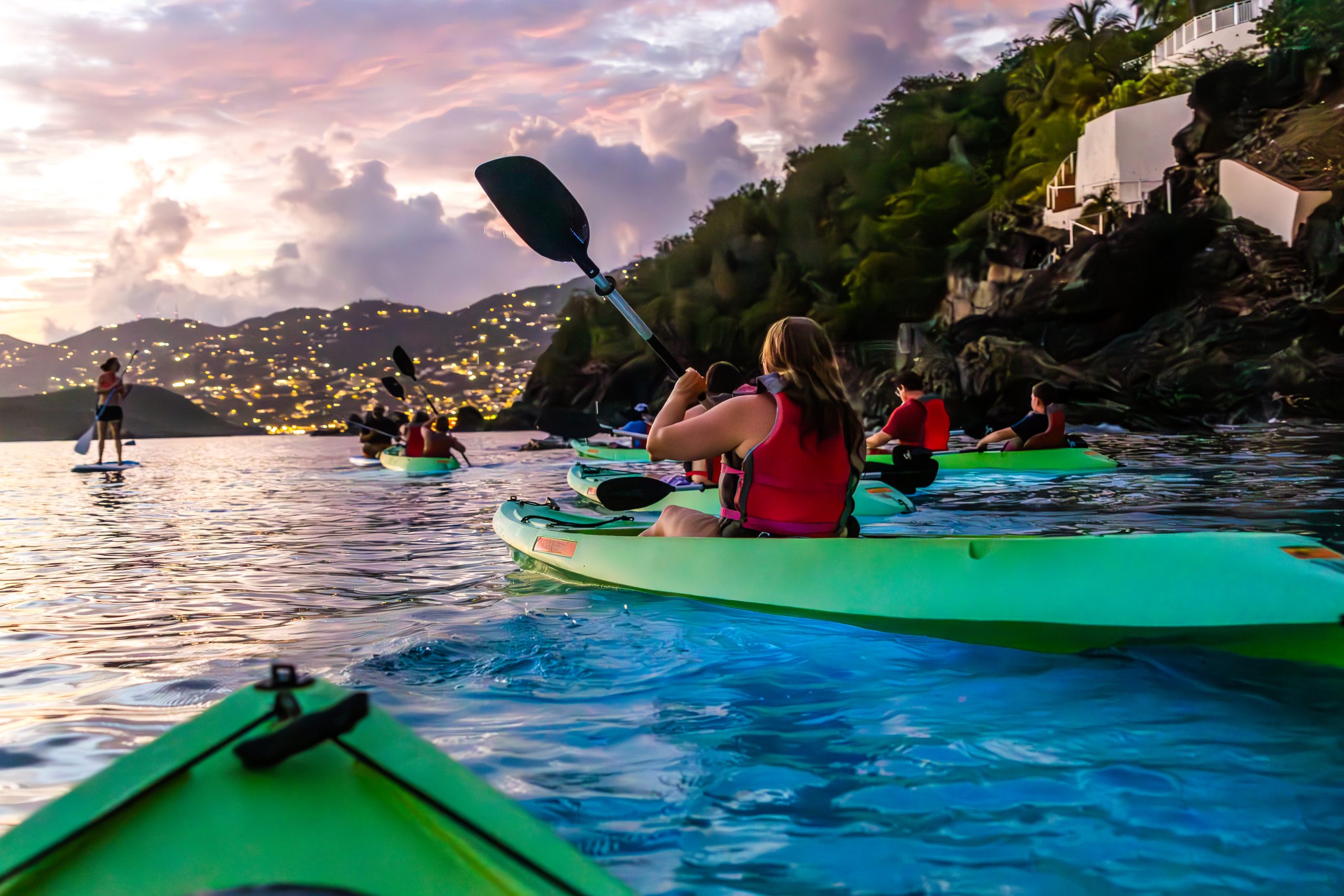 People kayaking on a lake at sunset with hill and buildings in background.