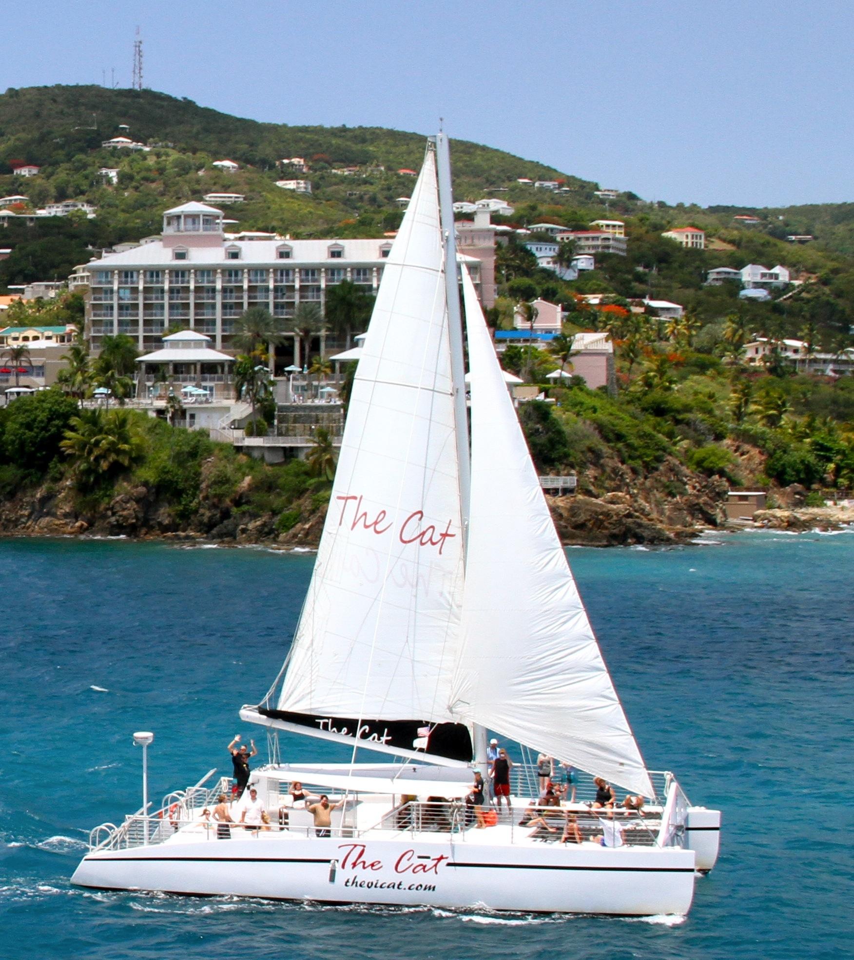 Sailboat 'The Cat' on blue water near a coastal town with green hills.