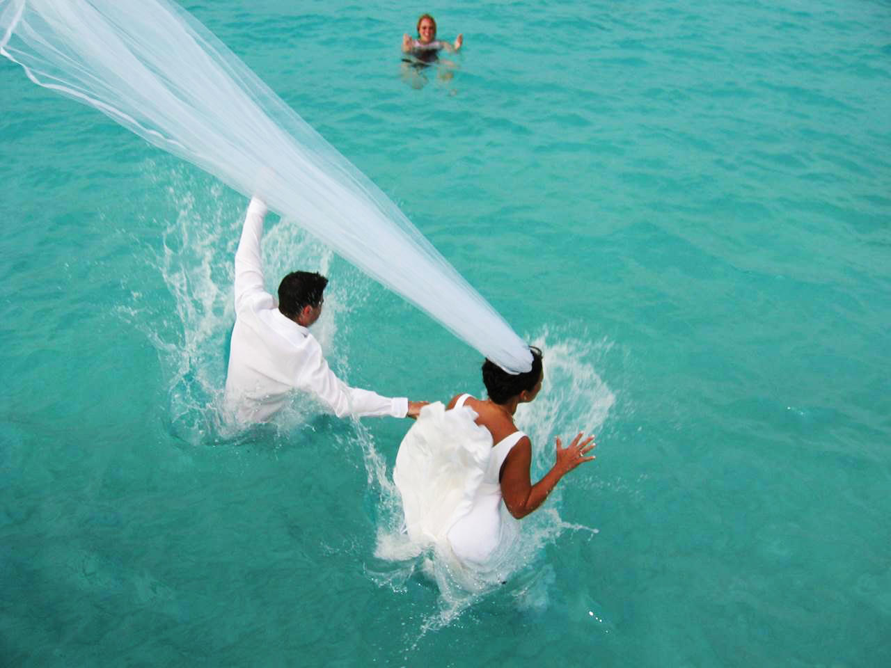 Bride and groom in water, bride's veil flowing, another person swimming.