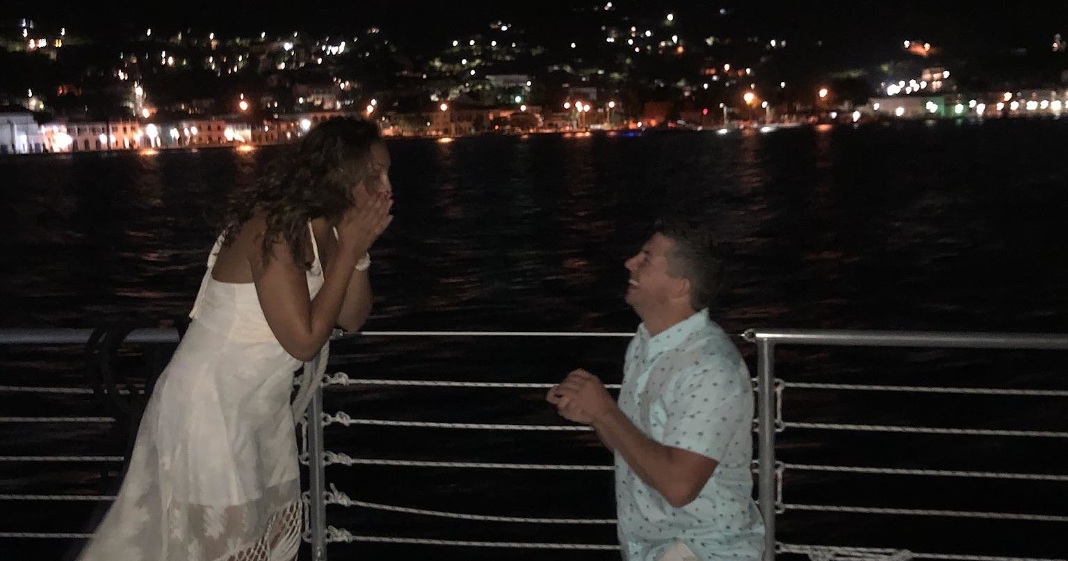 Man kneels proposing to woman on a boat at night, city lights in the background.