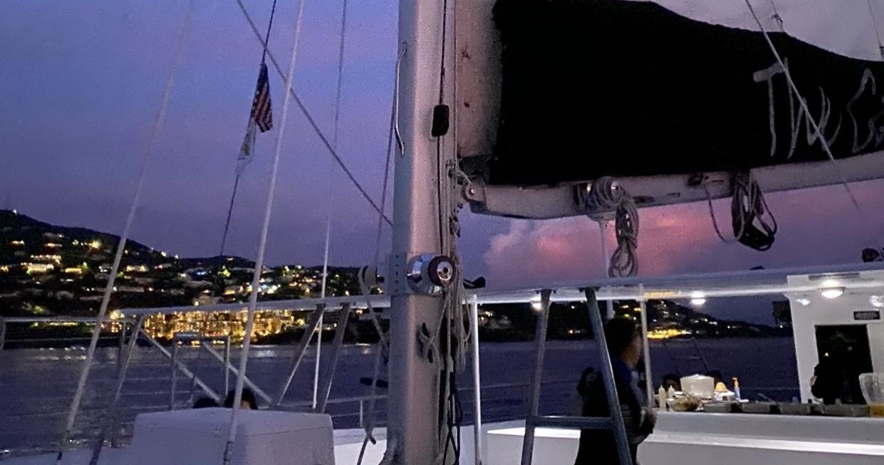 Sailboat deck at dusk with distant lit-up shoreline and hills.