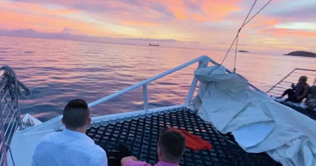Sunset view from a boat's deck with two people looking at the sea.