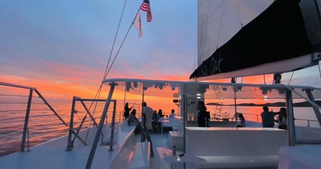 People on a boat deck at sunset with flags and sails visible.