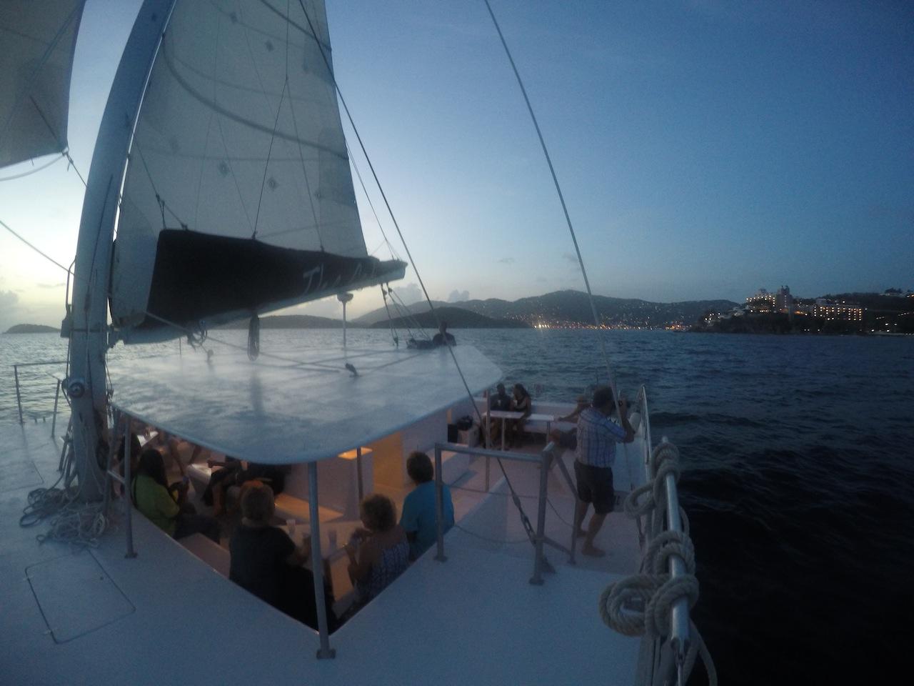 Sailboat at sunset with people on deck and distant shoreline view.