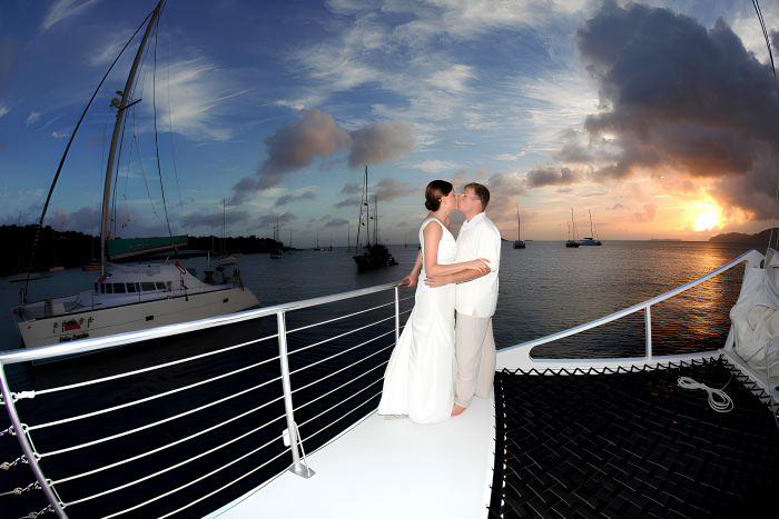 Couple in white clothing kiss on a boat deck at sunset with sailboats in the background.