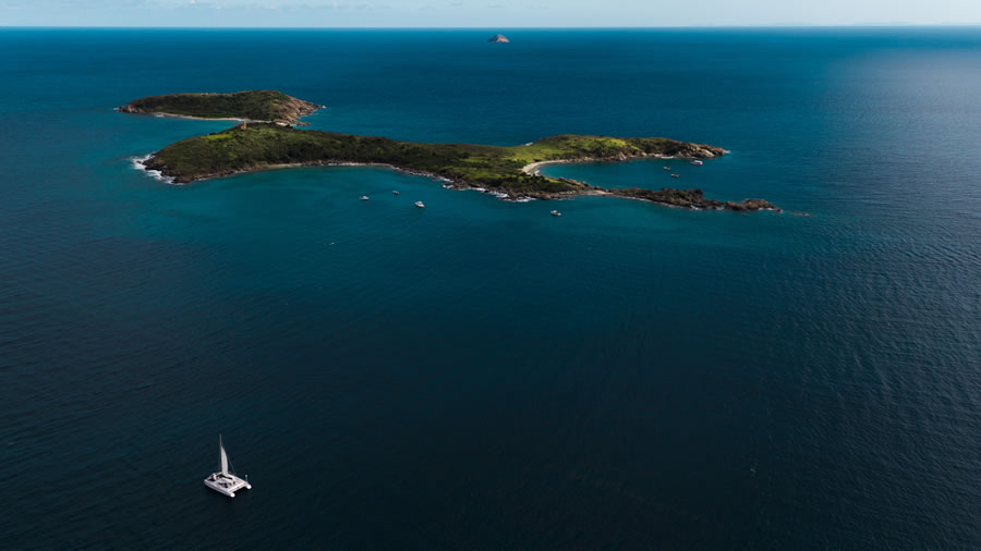 Aerial view of a sailboat near small green islands in a vast blue sea.