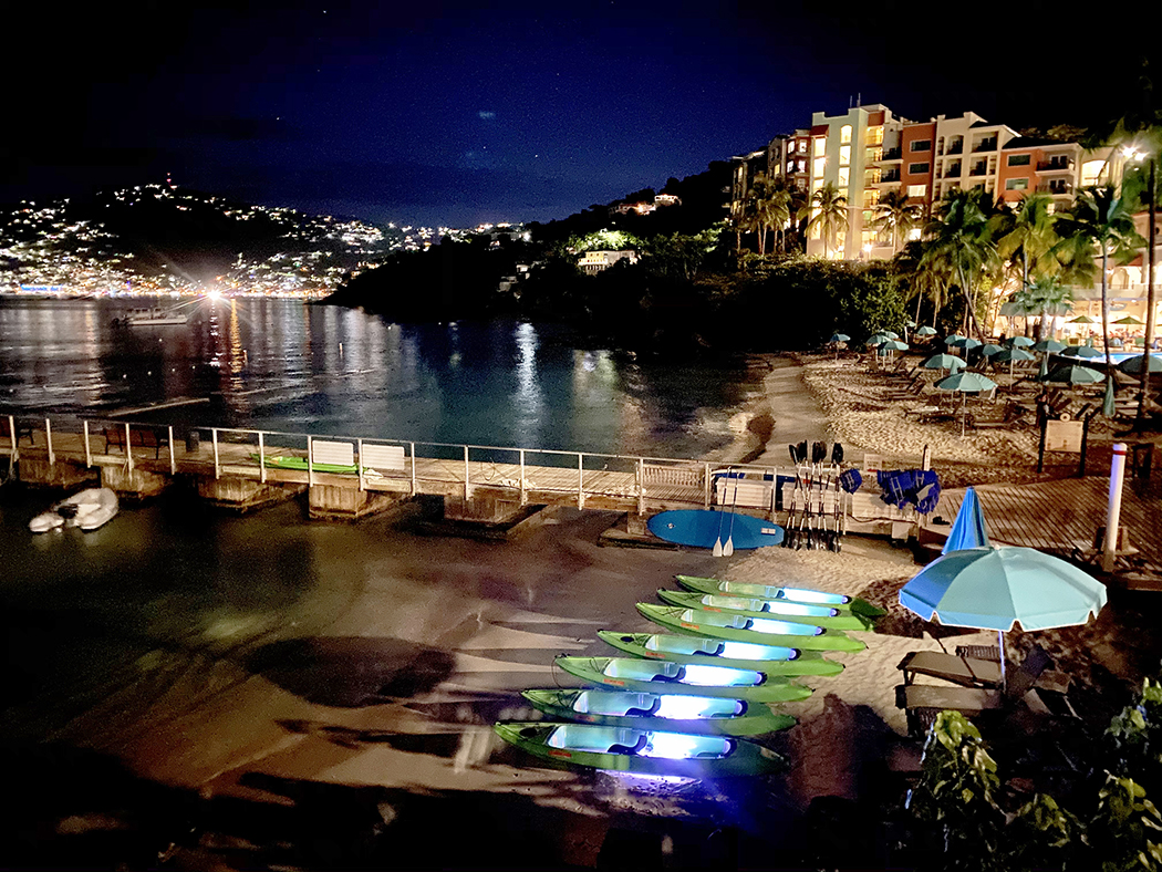 Night beach scene with lit kayaks, calm water, and city lights.